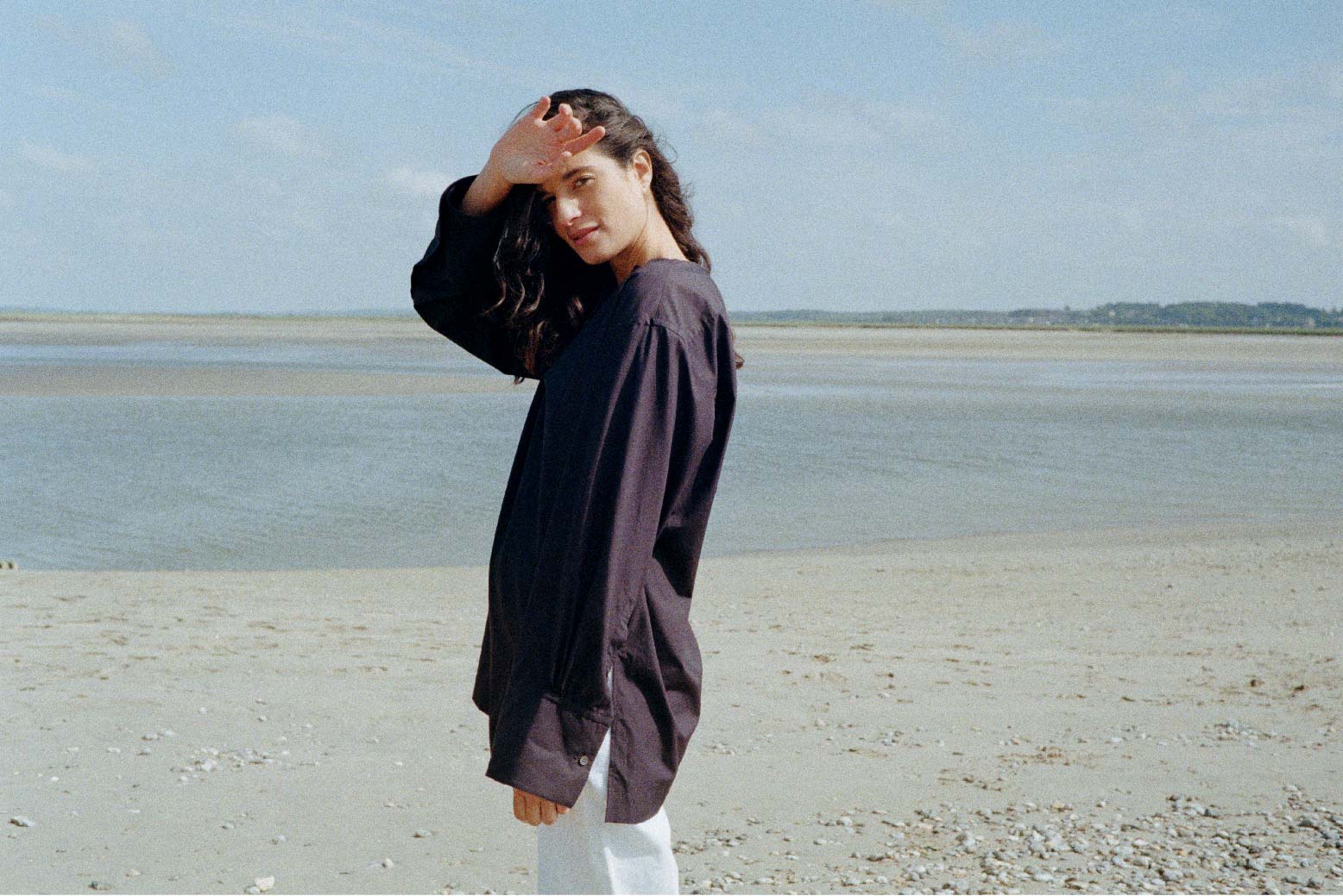 BOURRIENNE PARIS X: Photo vue de profil, le mannequin est sur la plage et porte une chemise bleu marine coupée dans une popeline de coton douce et de grande qualité d’origine japonaise. Le chemisier est non boutonné et a un décolleté fendu est orné de part et d'autre d'un plastron en ogive qui allonge la silhouette.