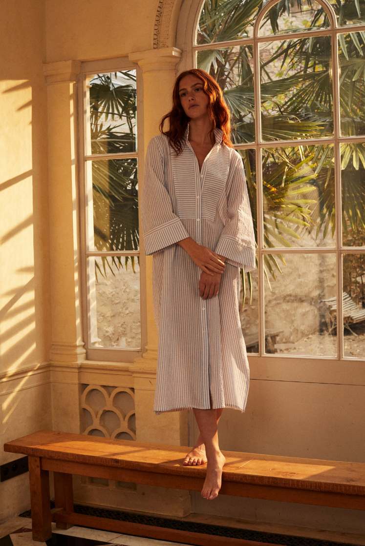 Photo plein pied d'une femme femme rouse debout sur un banc en bois. Elle porte une robe chemise ROSEE, elle est coupée dans un tissu seersucker rayé bleu et blanc de haute qualité.
Elle s'inspire des détails emblématique de la Maison Bourrienne Paris X: un col officier haut, plastron et poignets reprenant les iconiques de la Maison. Ses fentes sur le côté accentuent une silhouette féminine.