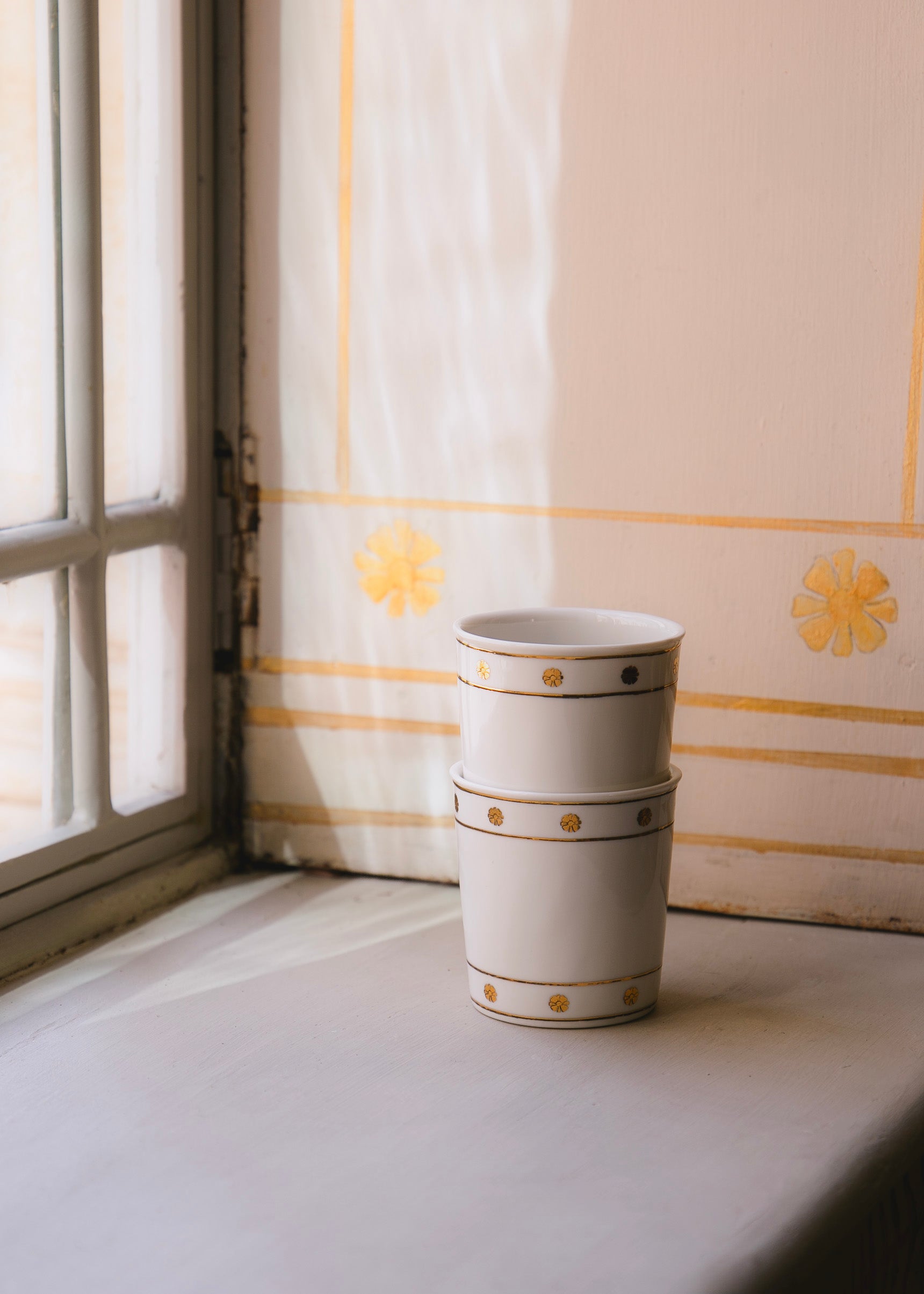 Deux timbales en porcelaine blanches ornées de motifs dorés empilées et posées dans un recoin de fenêtre. Le motif doré est inspiré des fresques de l'Hôtel de Bourrienne que l'on peut apercevoir derrière.