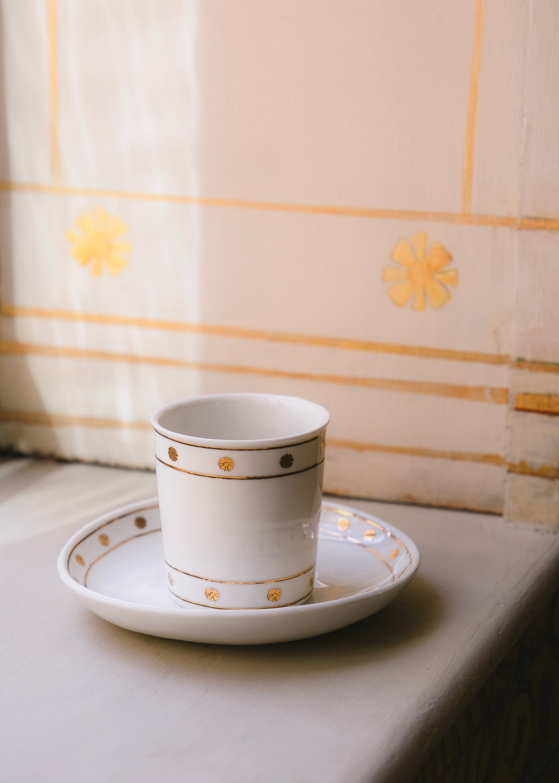 Timbale en porcelaine blanche ornée de motifs dorés posée dans la coupelle ornée du même motif. Le motif est inspiré des fresques de l'Hôtel de Bourrienne que l'on peut apercevoir en arrière plan.