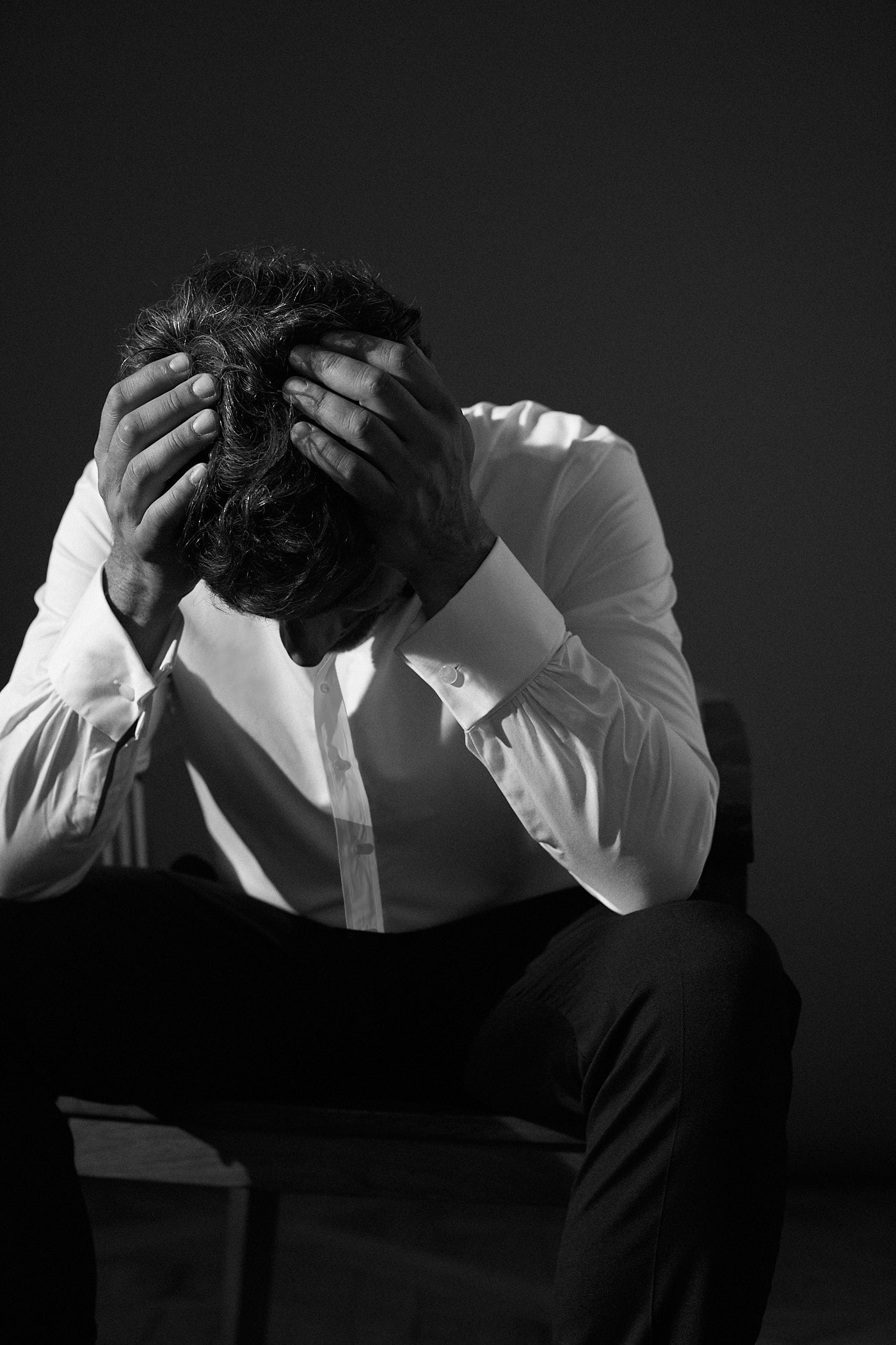 Photo en noir et blanc d'un homme assis sur une chaise qui baisse la tête et touche sa tête avec ses mains.On ne voit pas son visgae. Il porte une chemise blanche avec des poignets mousquetaires et un tailleur noir.