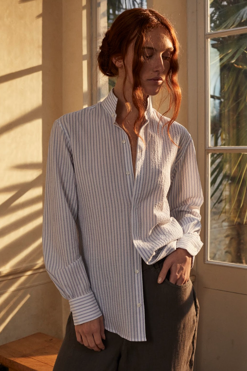 Femme portant la chemise AMAZONE en seersucker rayée bleue et blanche, dans un intérieur ensoleillé.