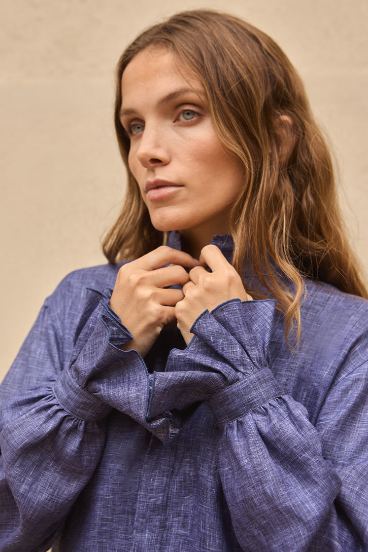 Femme portant une chemise en lin bleu avec des poignets à volants, tenant son col fermé.