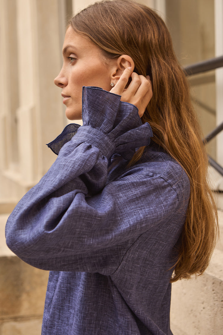 Femme portant une chemise en lin bleu avec des poignets à volants, remettant en place ses cheveux.