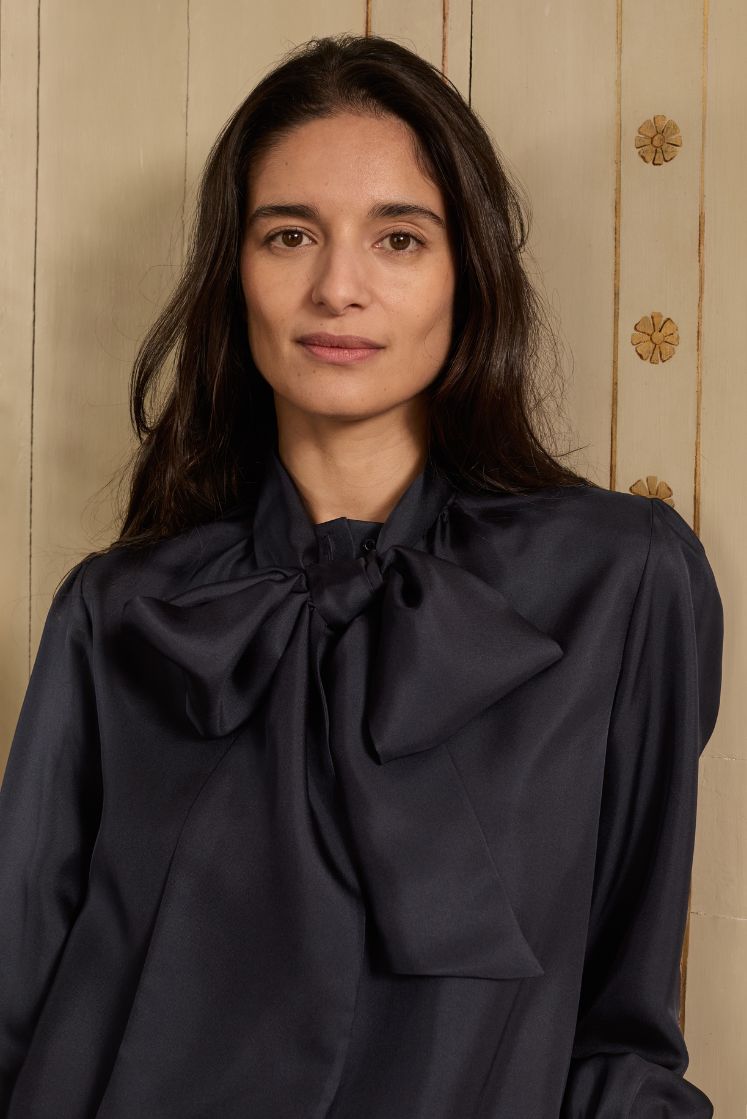 Femme brune portant la chemise COMTESSE en soie bleu marine avec le col lavallière attaché en noeud papillon.