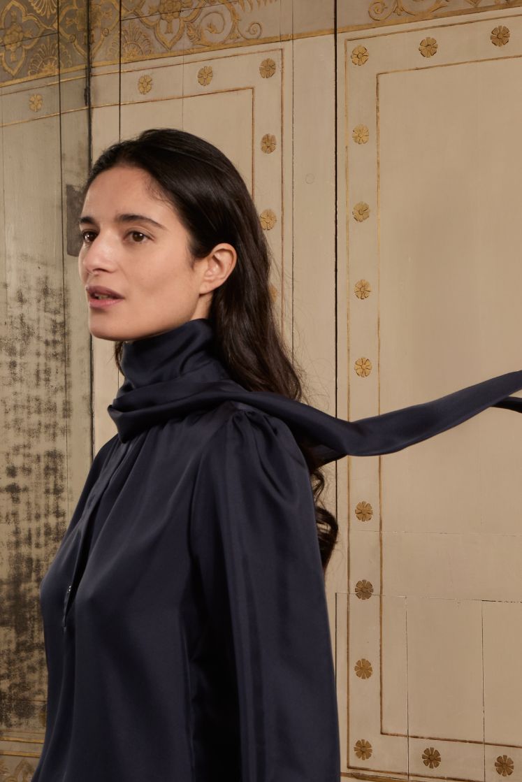 Femme brune portant la chemise COMTESSE en soie bleu marine avec le col lavallière enroulé autour du cou.