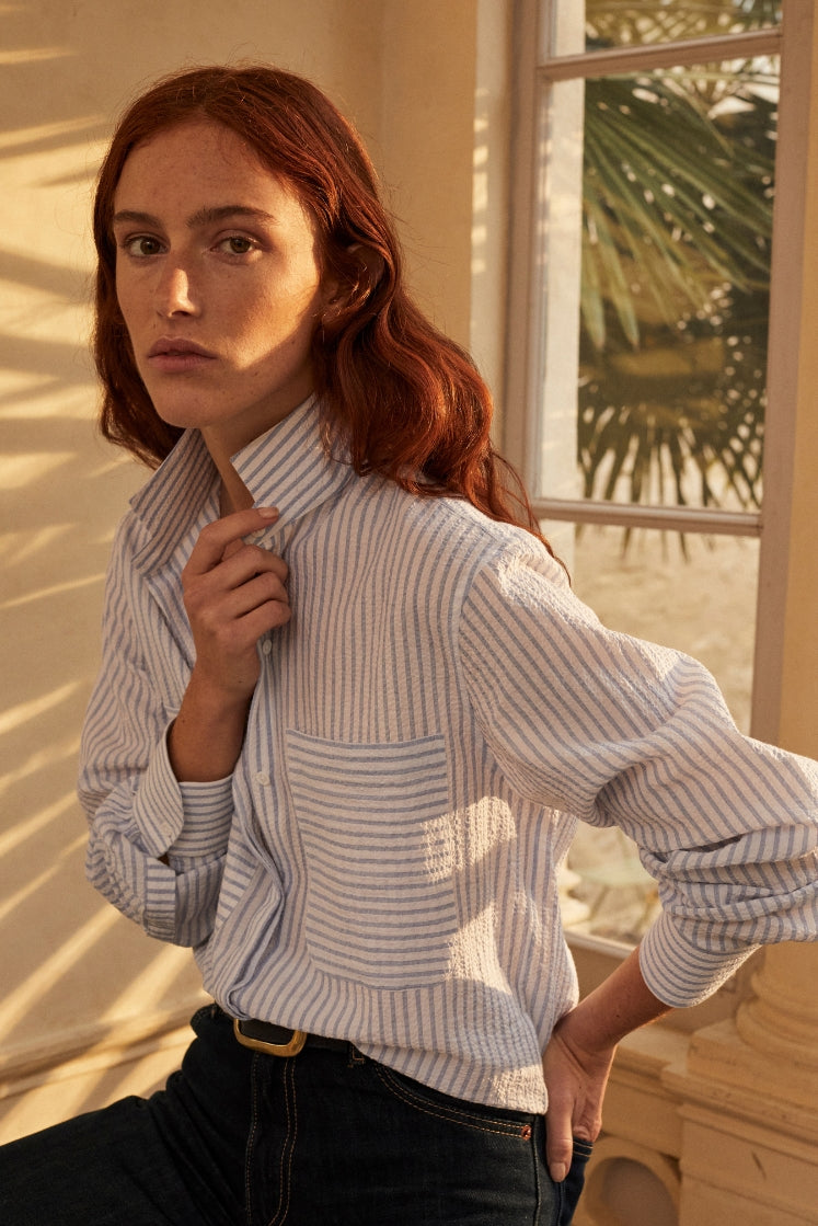 Une photo plan américain d'une femme rouse assise. Elle porte une chemise ENCRE SEERSUCKER un tissu rayé bleu et blanc. Elle a un col haut et des fronces aux manches, deux grandes poches sur le devant. La femme tient son col par la main droite, l'autre est à moitié rentrée sur sa poche arrière de jean.