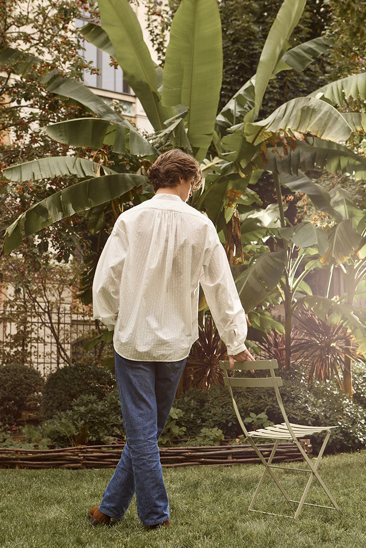 chemise blanche ample pour homme avec rayures blanches portée par un mannequin dans un jardin, de dos on distingue les fronces à l'arrière de la chemise