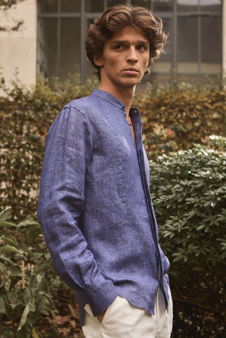 Homme dans un jardin portant une chemise en lin bleu au col officier, de profil.