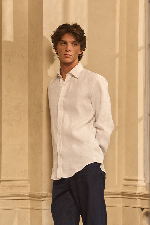 Homme portant une chemise blanche en lin, adossé à un mur, dans une lumière douce.