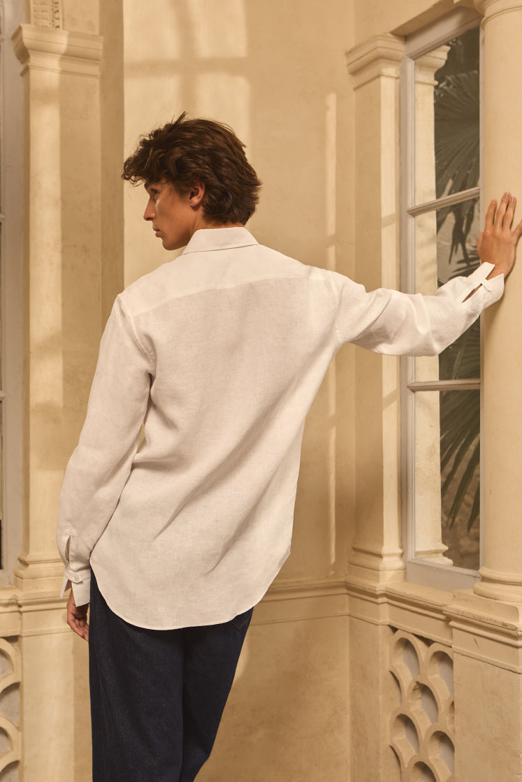 Homme portant une chemise blanche en lin, de dos, prenant appui sur un mur en pierre, dans un environnement ensoleillé.