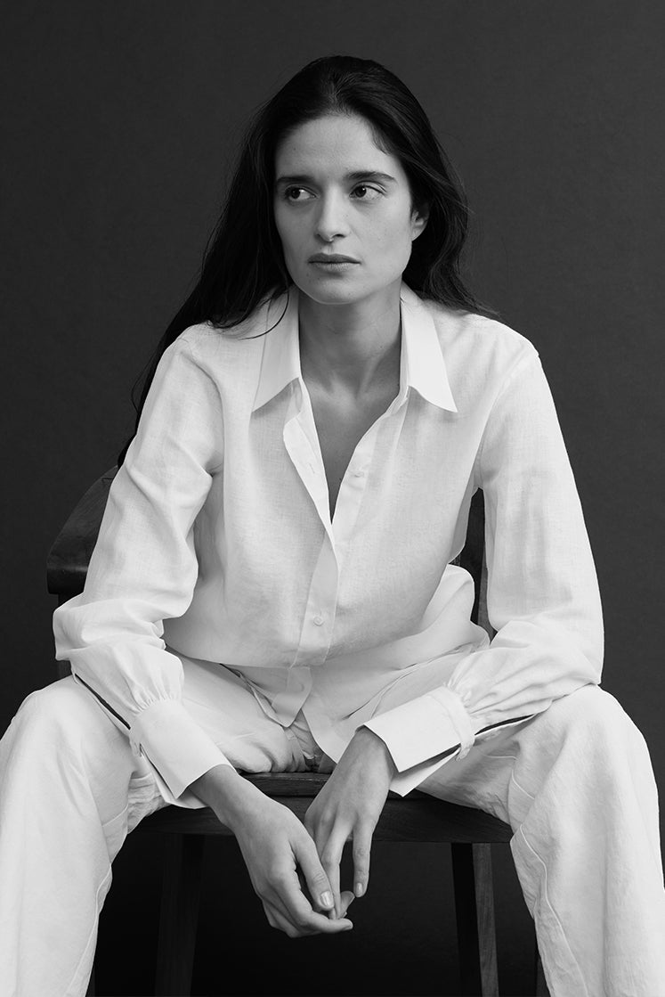 Femme brune assise sur une chaise portant une chemise en lin blanc à col français, légèrement déboutonnée, avec un pantalon en lin blanc.