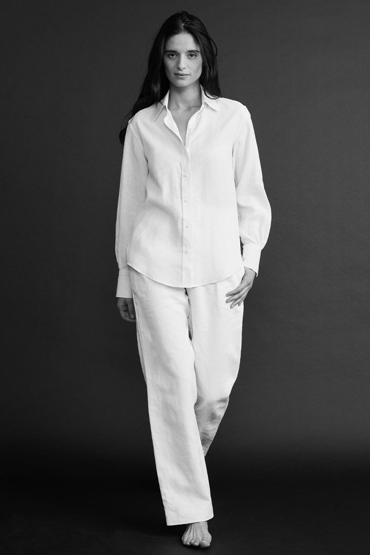 Plan plein pied d'une femme marchant, portant une chemise en lin blanc à col français, légèrement déboutonnée, avec un pantalon en lin blanc.