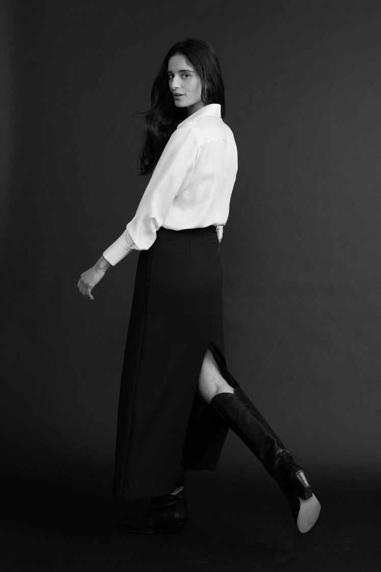 Femme portant la chemise NUMÉRO VII en soie blanche, avec une jupe longue noire et des bottes, prise de dos en marchant.