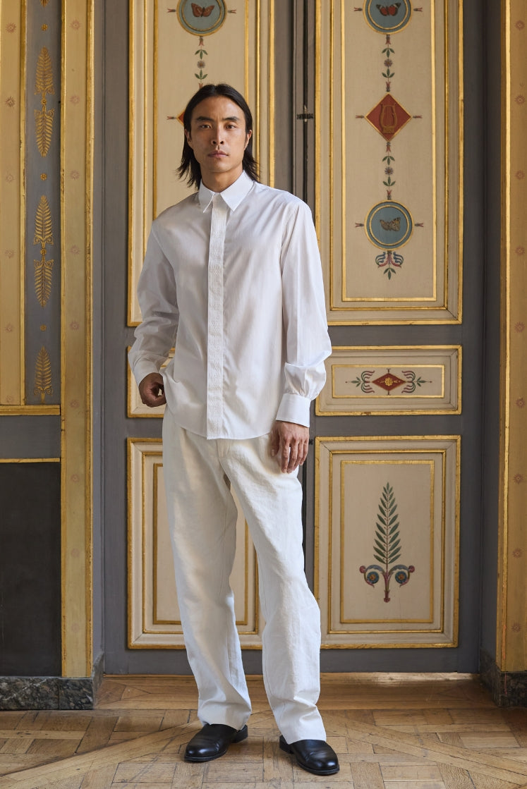 Homme portant la chemise ESQUISSE avec son col français et son galon brodé aux motifs de l'Hôtel le long de la ligne de boutonnage, avec le pantalon HORIZON en lin plâtré.
