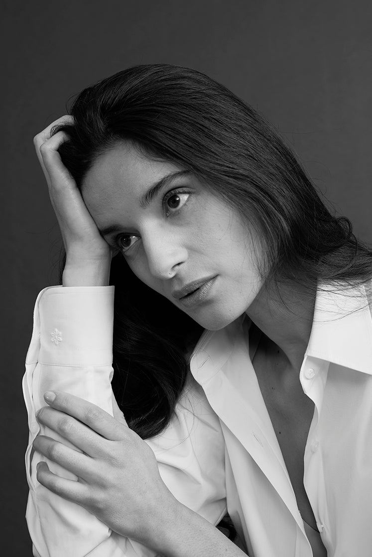 Portrait en noir et blanc d’une femme aux cheveux longs portant une chemise blanche à poignet classique brodée de la Fleur de Bourrienne, la tête appuyée sur sa main, regard pensif sur fond sombre.