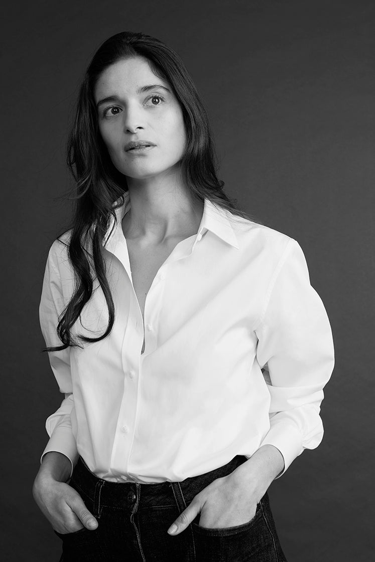 Portrait en noir et blanc d’une femme portant une chemise blanche et un jean sombre, les mains dans les poches, regard tourné vers le haut sur fond sombre.
