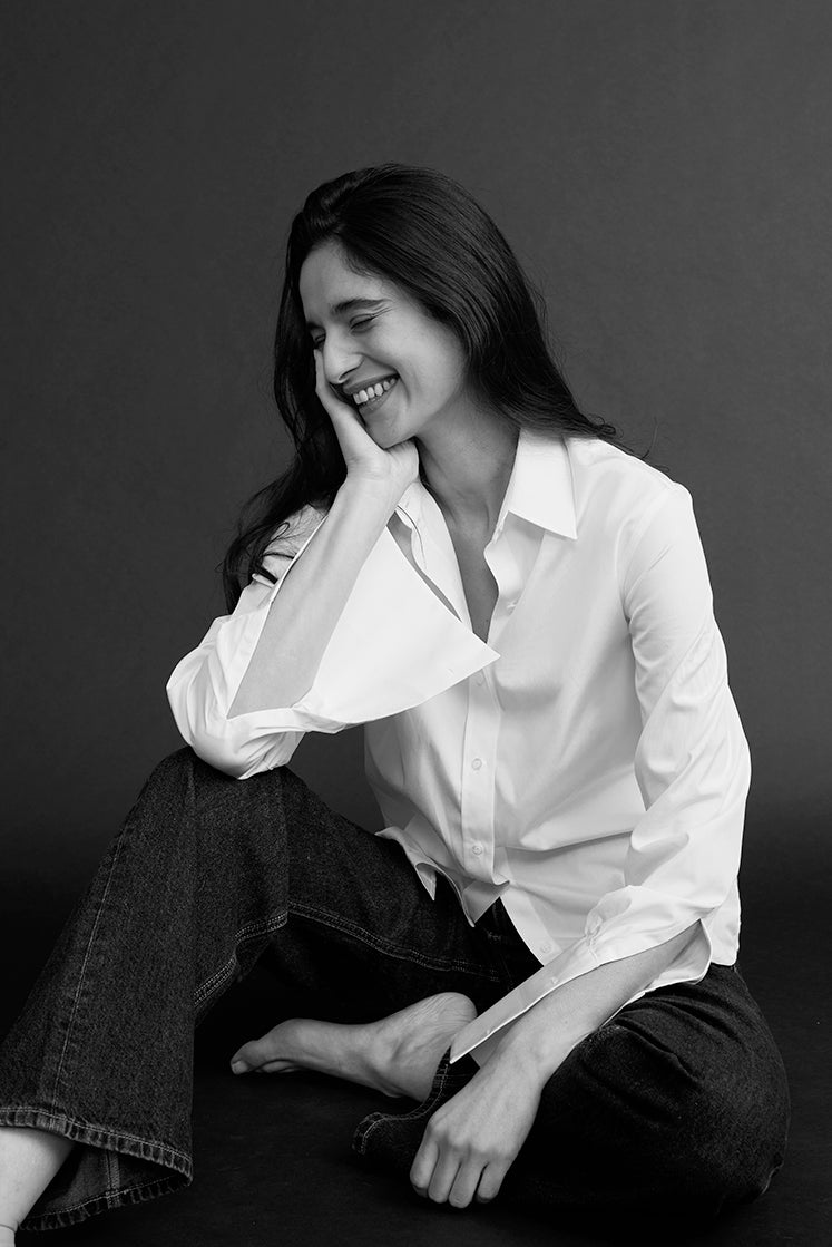 Portrait noir et blanc d’une femme souriante assise en chemise blanche et jean noir, à col français et poignets mousquetaires détachés.