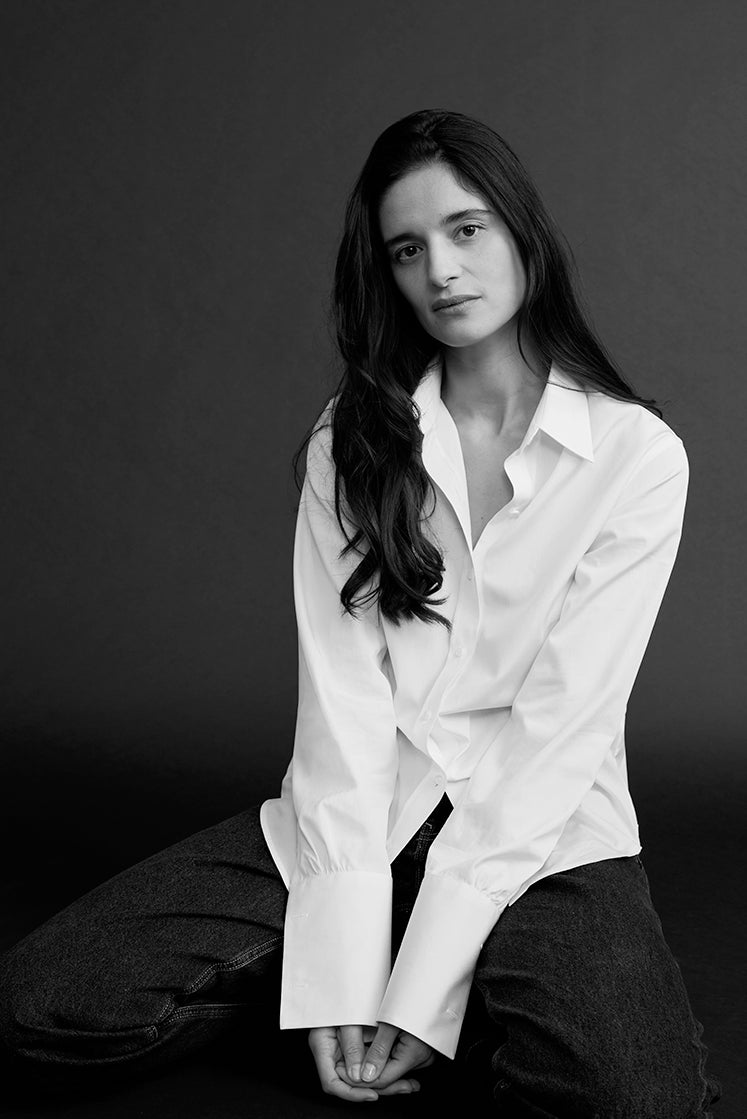 Portrait noir et blanc d’une femme assise en chemise blanche intemporelle, à col français et poignets mousquetaires détachés.