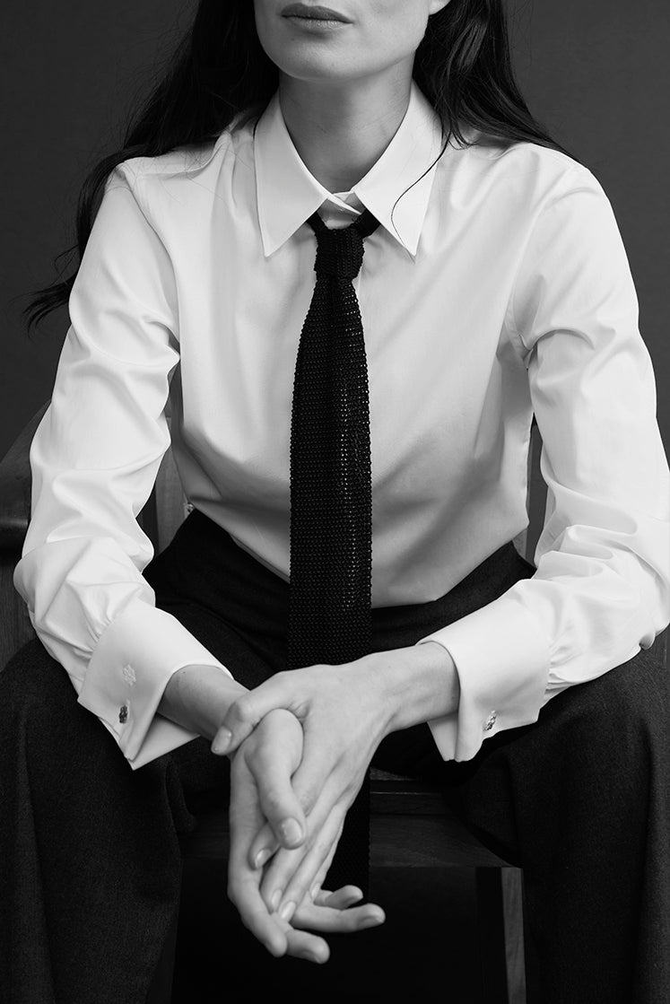 Portrait noir et blanc d’une femme assise sur une chaise en chemise blanche à col français stylisée avec une cravate noire.