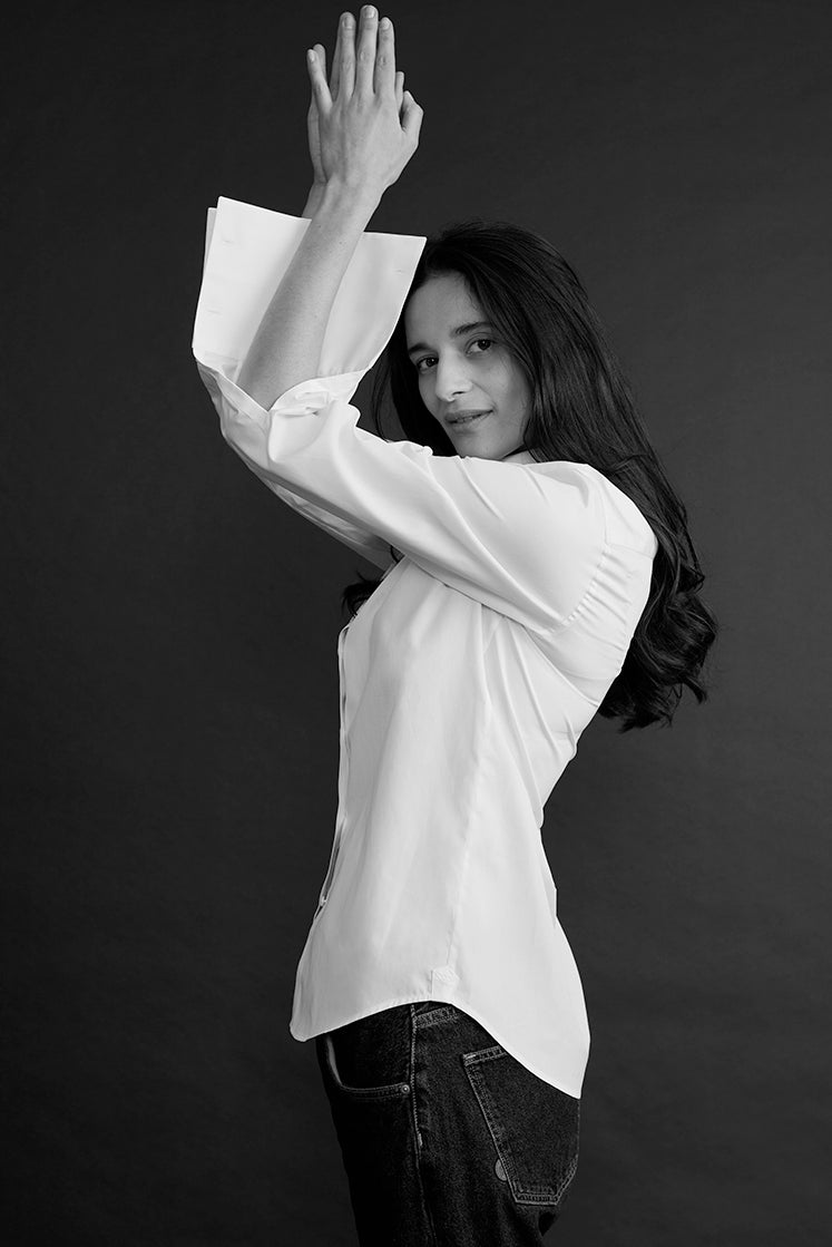 Portrait en noir et blanc d’une femme portant une chemise blanche et un jean sombre, posant de profil avec les bras levés au-dessus de la tête sur un fond sombre.