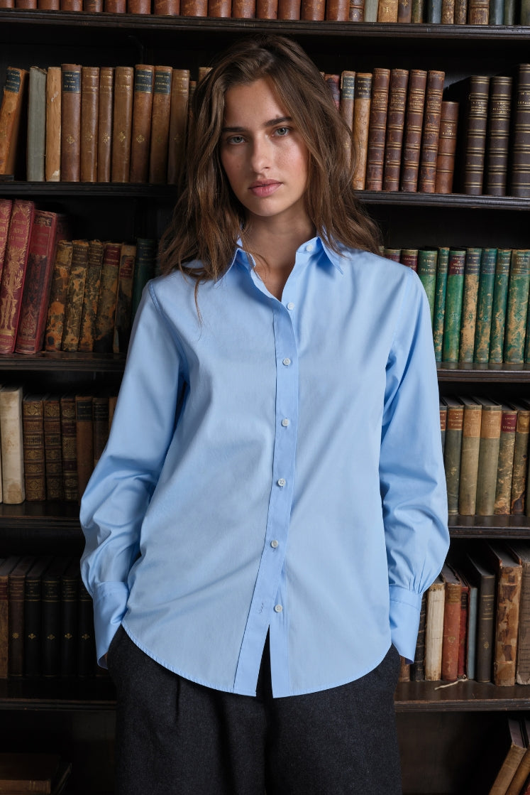 Femme dans une bibliothèque portant une chemise bleu ciel en popeline de coton à col français.