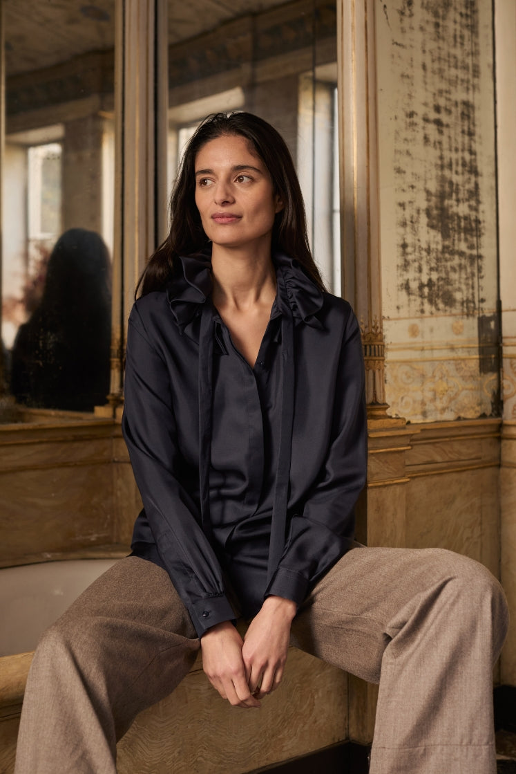 Mannequin assise dans une salle de bain historique, portant la chemise PASTEL en soie navy avec son col à volants.