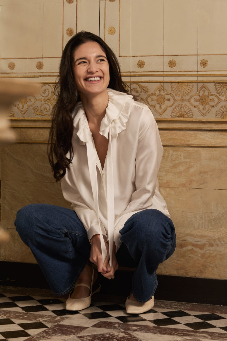 Mannequin accroupie dans la salle de bain de l'Hôtel de Bourrienne, portant la chemise PASTEL en soie blanche avec son col à volants, un jean bleu et des ballerines blanches.