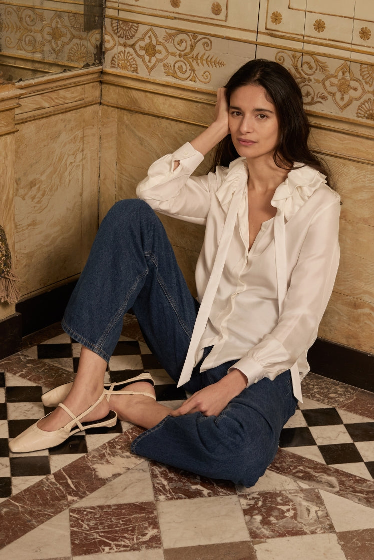 Mannequin assise sur le sol à carreaux de la salle de bain de l'Hôtel de Bourrienne, portant la chemise PASTEL en soie blanche avec son col à volants, un jean bleu et des ballerines blanches.