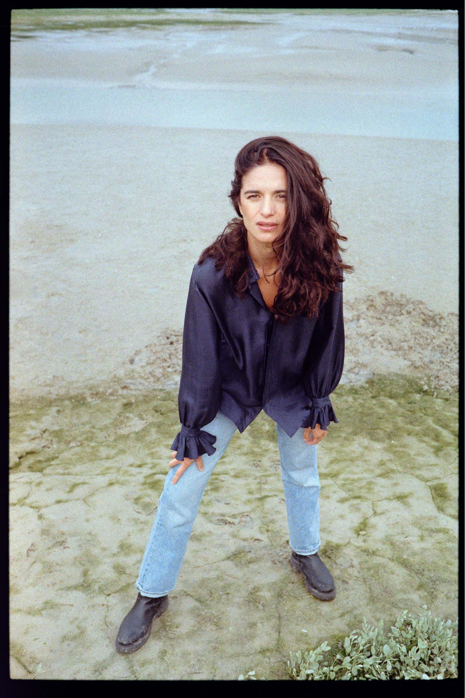 BOURRIENNE PARIS X: Photo vue de face et prise de haut du mannequin, elle est sur la plage et porte une chemise en soie bleu marine, on aperçoit les détails des manches bouffantes et des volants brodés. 