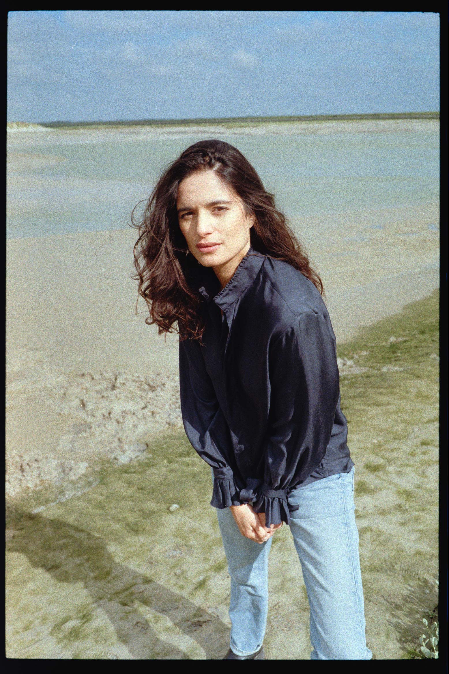 BOURRIENNE PARIS X: Photo vue de profil, le mannequin est sur la plage et porte une chemise en soie bleu marine, on aperçoit les détails du col officier haut, rebrodé d’un galon, des manches bouffantes et de leurs volants brodés ainsi que des fronces qui se situent dans le dos de la chemise. 