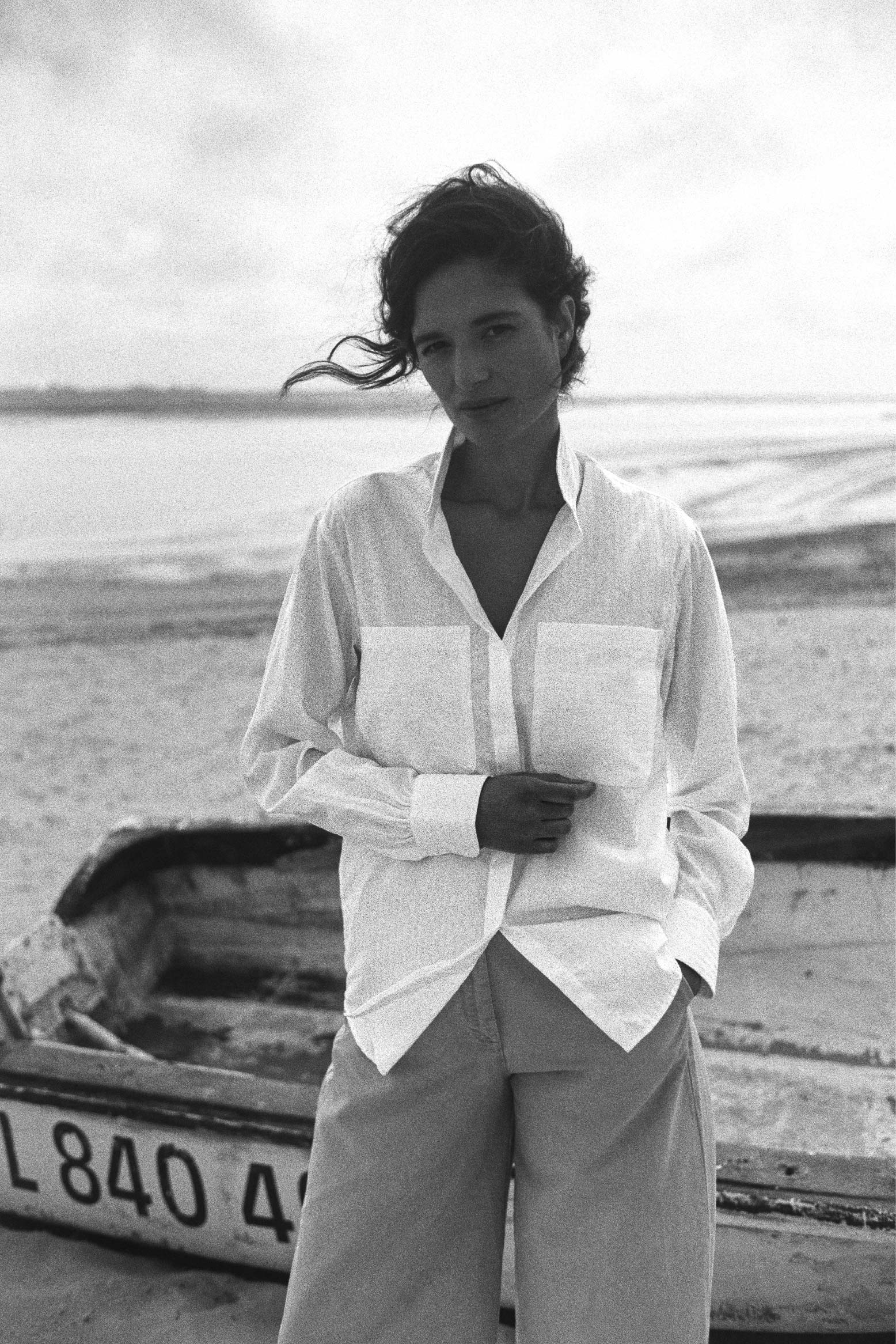 BOURRIENNE PARIS X: Photo sur la plage et vue de face, le mannequin porte une chemise en coton et à rayures blanches ton sur ton, chemise à col haut et à poches plaquées sur le buste, la photo est en noir et blanc.