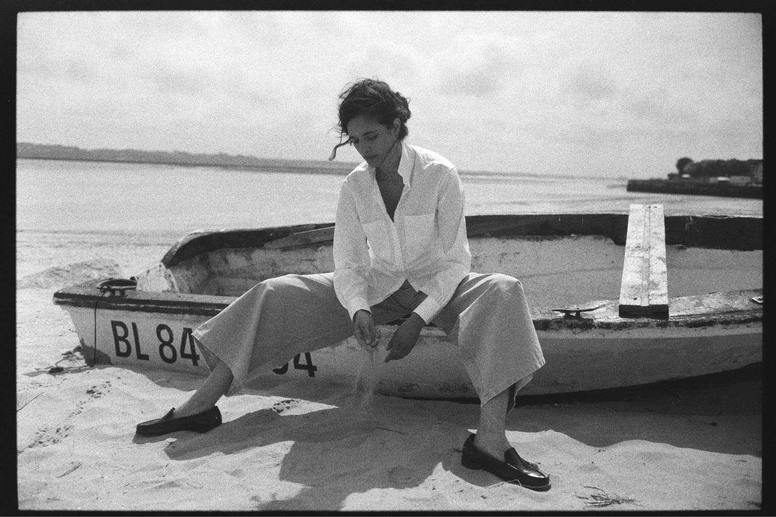 BOURRIENNE PARIS X: Photo vue de face, le mannequin est assise sur une épave d’un bateau sur la plage, elle porte une chemise en coton et à rayures blanches ton sur ton. 