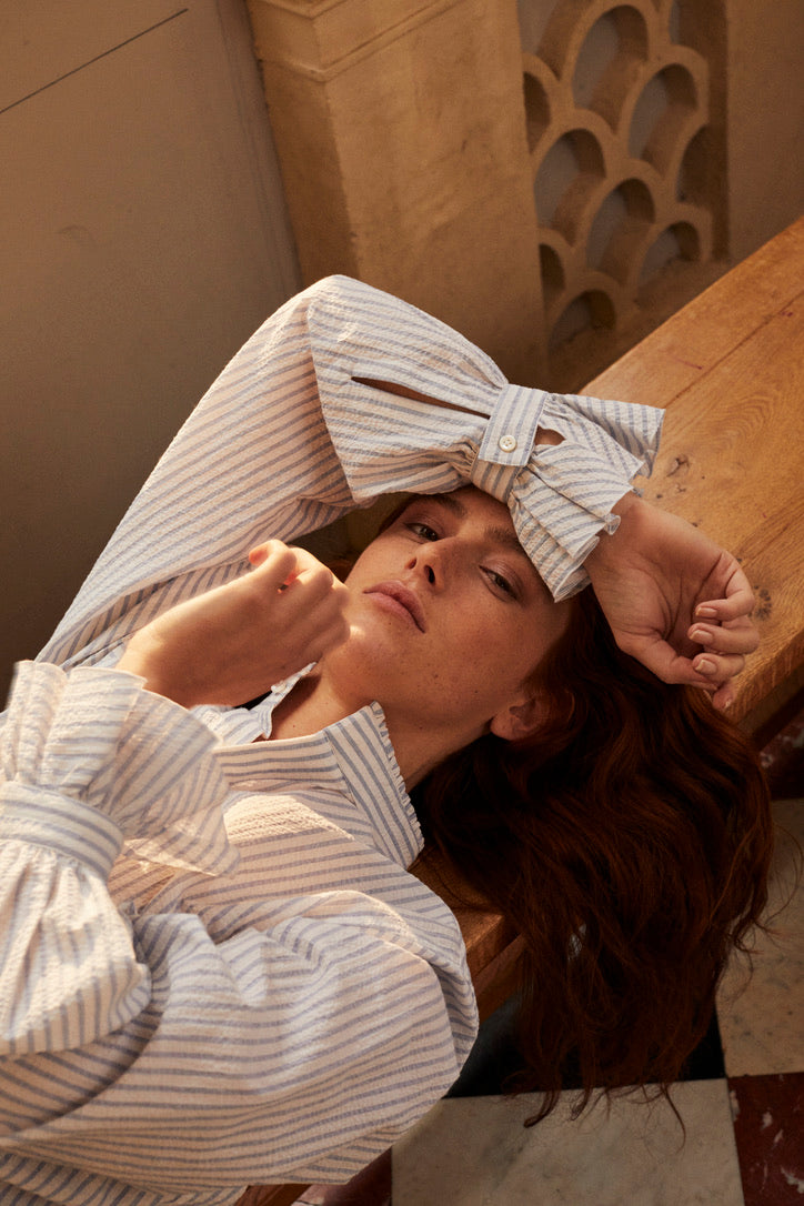 Photo plan serré en plongée, d'une femme rousse allongée sur un banc en bois.Elle porte une chemise Boudoir Seersucker Rayée Bleu qui est rentrée sous un jean taille haute. Son avant bras est posé sur son front, nous apercevons plus en détails les volants des poignets, signature de la Maison Bourrienne. 