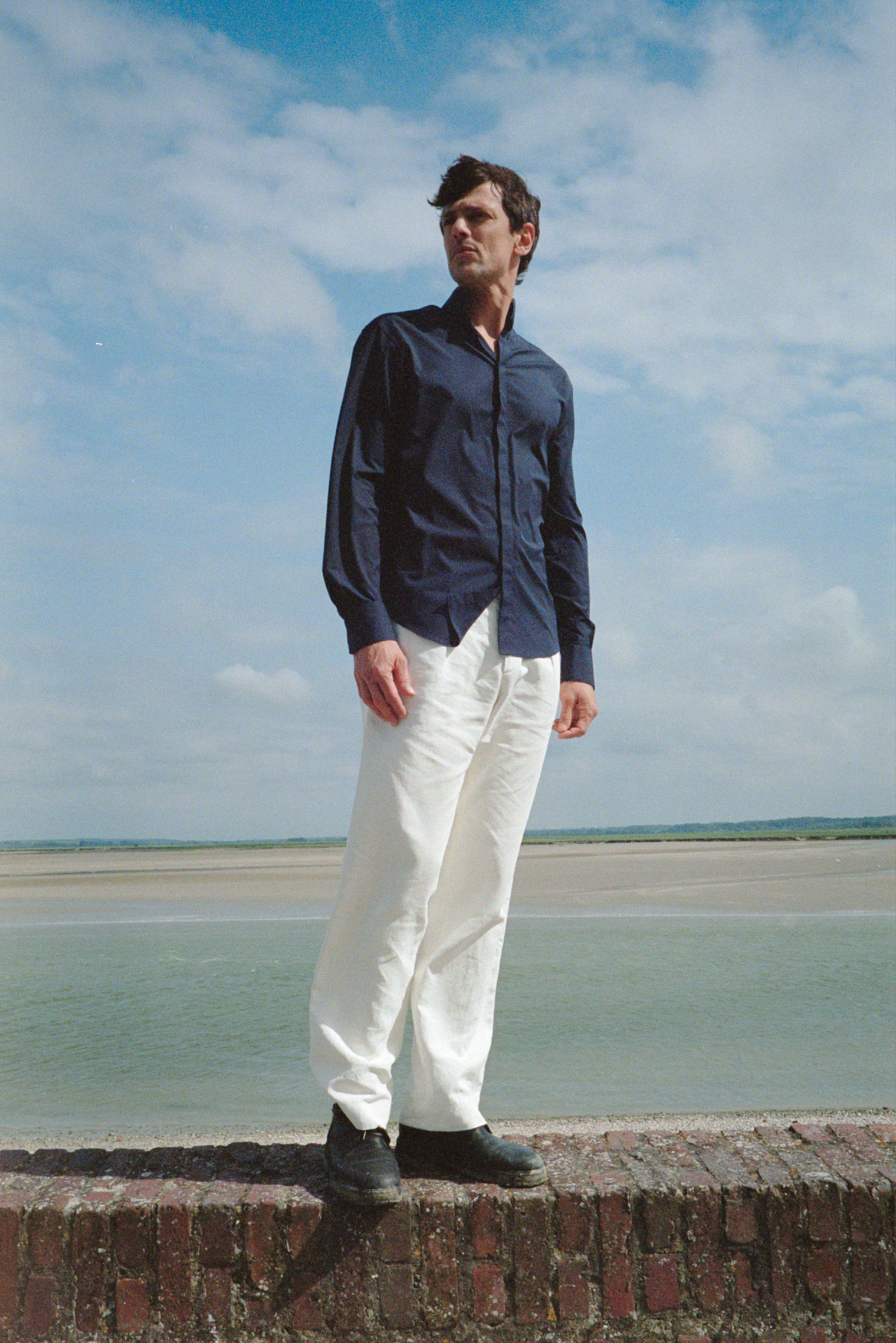 BOURRIENNE PARIS X: Photo prise de plain pied, le mannequin est sur un muret, dos à la mer, regarde à l’horizon et porte une chemise en popeline bleu marine. Le col de la chemise est haut, et les poignets classiques avec des fronces. 