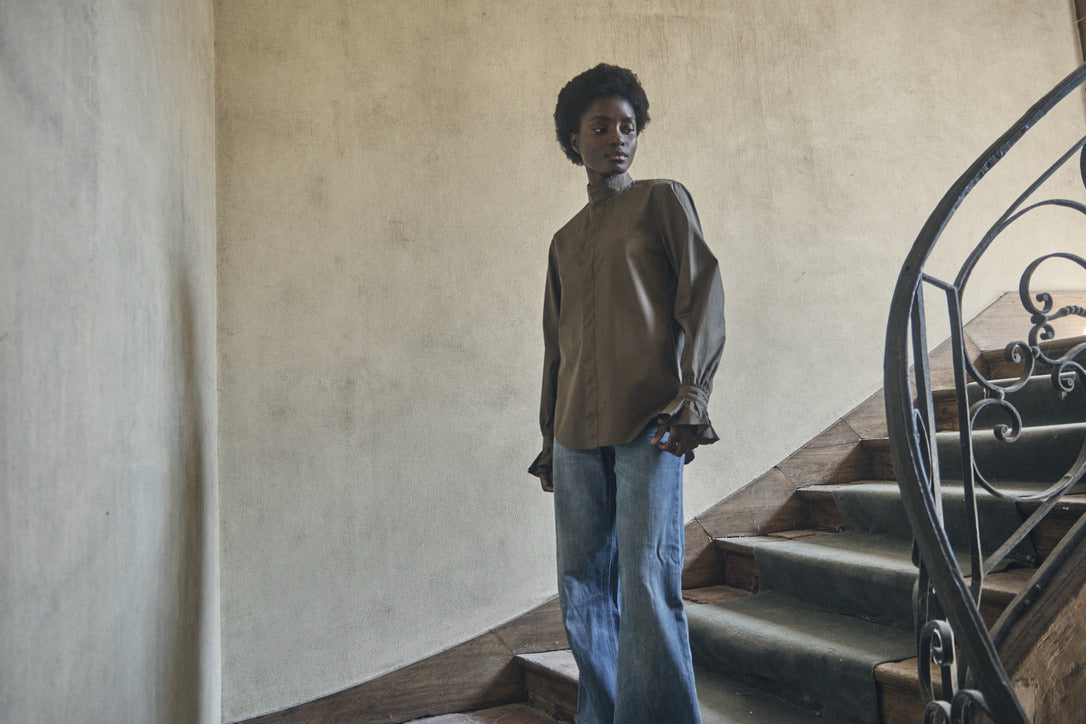 Femme portant la chemise BOUDOIR couleur Kaki et un jean bleu, posant dans les escaliers d'une maison, prise en contre-plongée du bas des escaliers.