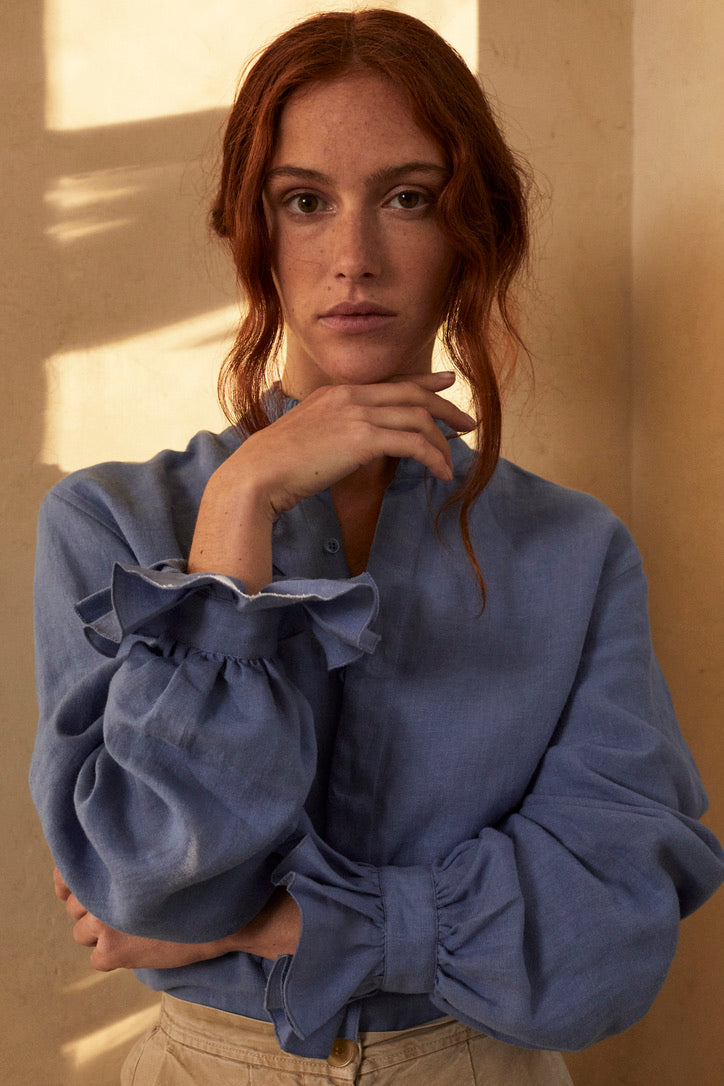 Femme rousse portant une chemise en lin bleu BOUDOIR de Bourrienne Paris X, avec col ancien et poignets à volants. Elle pose dans une lumière dorée, projetant des ombres élégantes sur le mur.