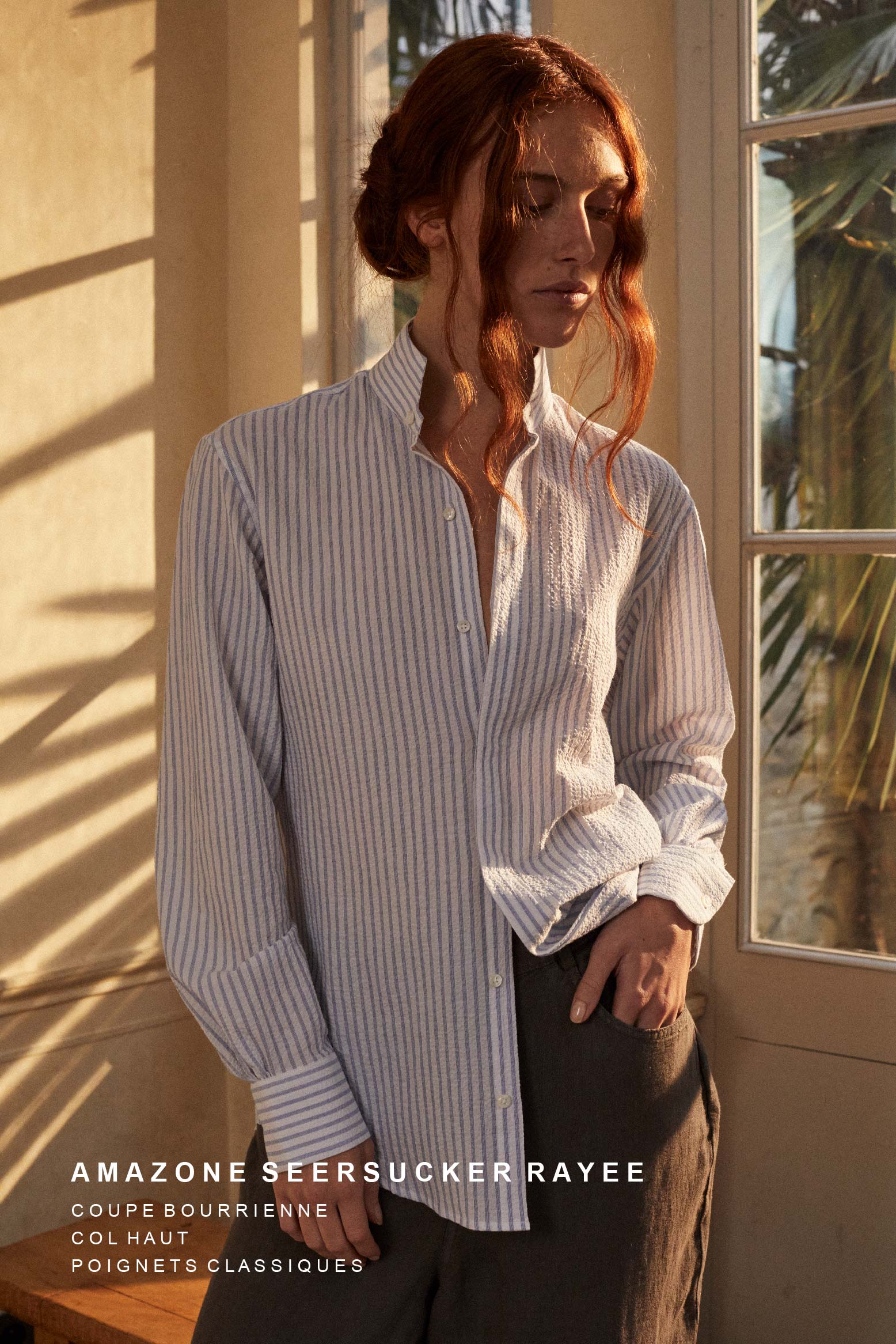 Photo plan américain d'une femme rousse  debout, portant un chignon, deux mèches  tombent sur son visage. Elle porte une chemise AMAZONE SEERSUCKER dans un tissu rayé bleu et blanc en seersucker. La chemise emprunte son col ancien au vestiaire masculin, l'adaptant avec subtilité à la femme. Son volume est sublimé par une série de fronces dans le dos. 