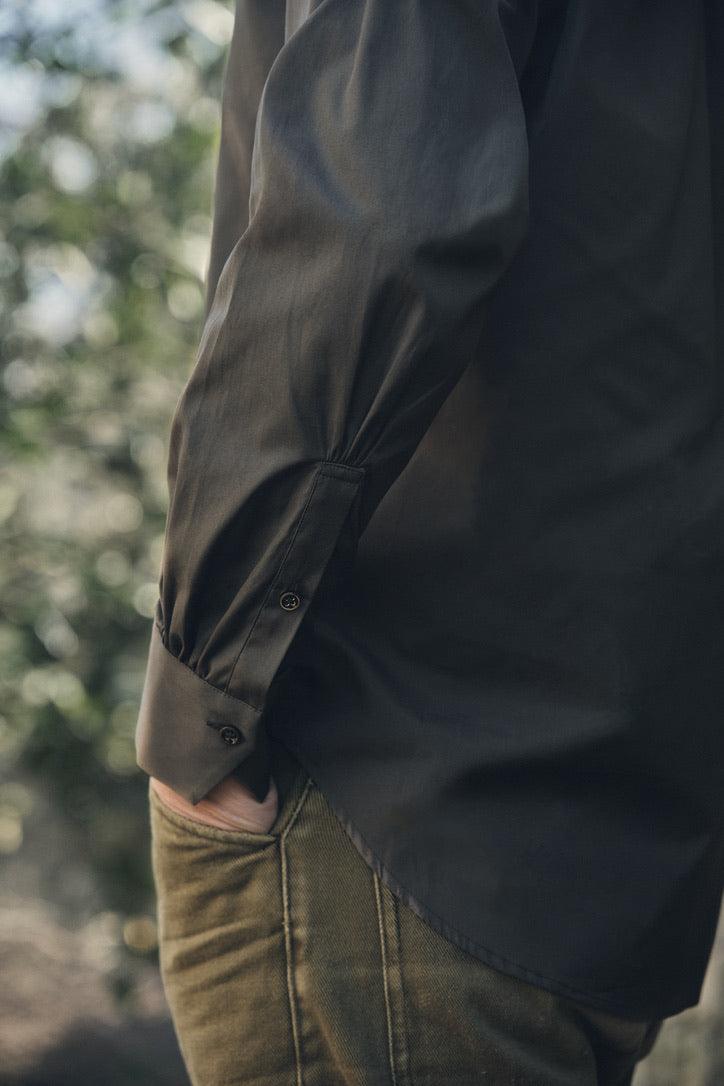 Détail de la chemise ÉPICURIEN Kaki en popeline de coton : vue du bas de manche ample finie par un poignet classique et un pli discret, glissant sur un pantalon kaki, le tout dans une lumière naturelle et un arrière-plan végétal flou.