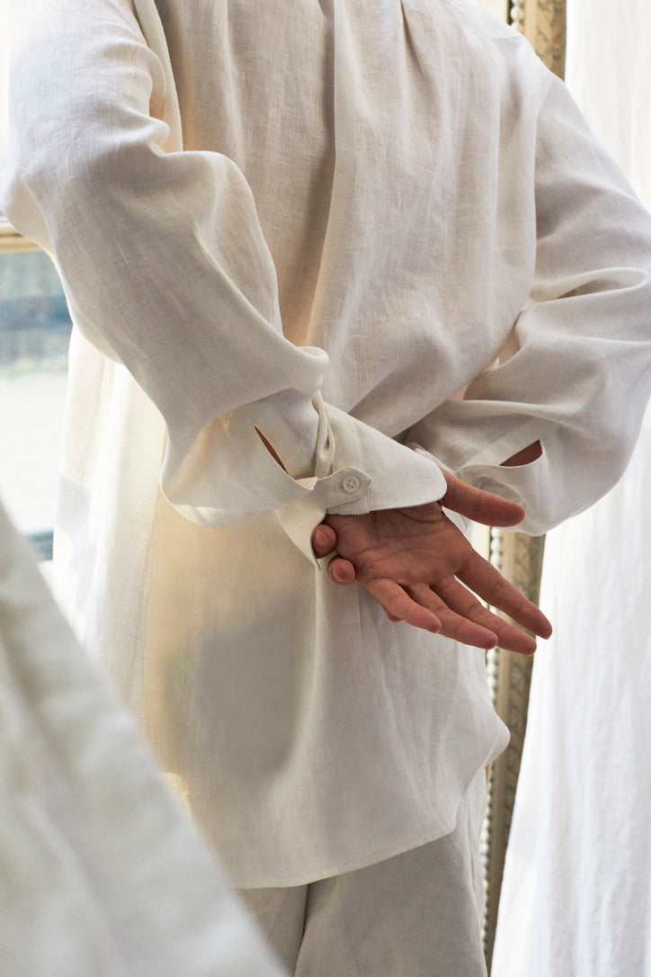 Photo des poignets Bourrienne signature de la Maison. Le mannequin est de dos et se tient les mains. la chemise est en lin léger. on discerne des rideaux de lin autour de lui légèrement floutés.