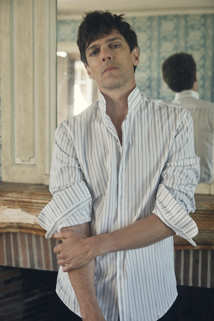 Un homme debout face à l’objectif, les manches de sa chemise retroussées au-dessus des coudes et la main droite saisissant l’avant-bras gauche, arbore une allure à la fois naturelle et assurée. Sa chemise ajustée en popeline kaki clair, parcourue de fines rayures verticales blanches et gris clair, se ferme par un col montant haut et ouvert jusqu’au deuxième bouton. Le tissu fluide forme de légers plis sur le torse, soulignant la coupe près du corps et la matière.