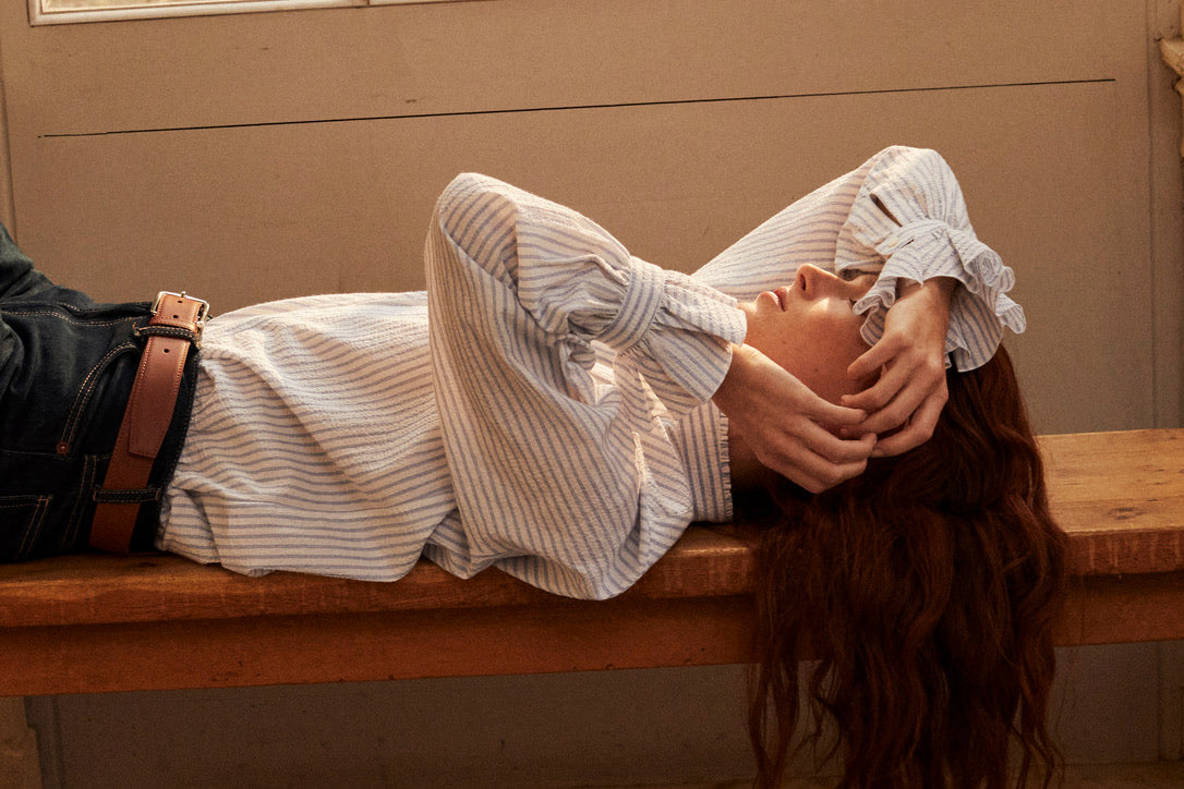 Photo plan serré, d'une femme rousse allongée sur un banc en bois. Elle est de profil, elle porte une chemise Boudoir Seersucker Rayée Bleu qui est rentrée sous un jean taille haute. Son avant bras est posé sur son front, nous apercevons plus en détails les volants des poignets, signature de la Maison Bourrienne. 