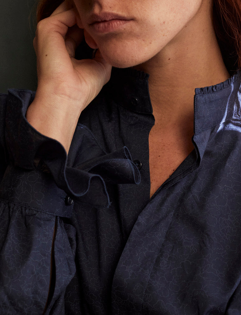 Photo gros plan d'une femme qui porte une chemise BOUDOIR bleu marine imprimé liberty de la marque Bourrienne Paris X. La femme rousse pose sa main gauche sur sa machoire. On aperçoit le col ancien de la chemise qui se fini d’un liseré de petits volants lui donnant un côté sophistiqué. Les manches bouffantes sont ornées d’un volant ce qui donne un côté original à la chemise. 