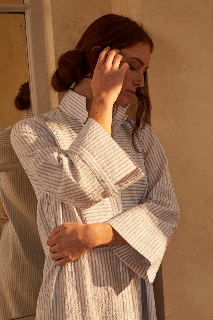 Photo plan américain d'une femme rousse portant un chignon, une mèche tombe sur son profil gauche. Elle porte une robe chemise ROSEE, elle est coupée dans un tissu seersucker rayé bleu et blanc de haute qualité. Elle a un col officier haut, plastron et poignets reprenant les iconiques de la Maison. Ses fentes sur le côté accentuent une silhouette féminine. Sa main droite touche délicatement son visage, on aperçoit les détails de la manche 3/4 et son avant-bras gauche entoure son ventre. 