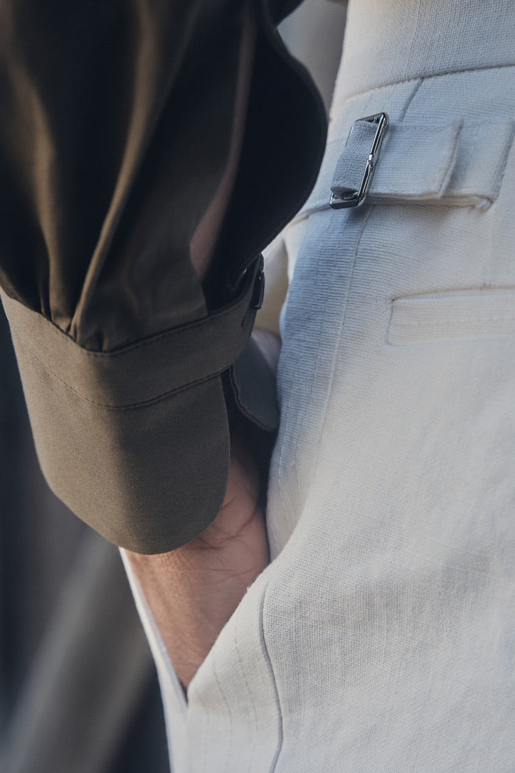 Gros plan sur le poignet droit glissé dans la poche d’un pantalon en lin plâtré blanc taille haute, révélant la manche droite retroussée de la chemise kaki foncé et son poignet droit fermé par un simple bracelet. Le réglage latéral du pantalon, orné d’une boucle métallique, contraste avec la texture satinée du tissu kaki.