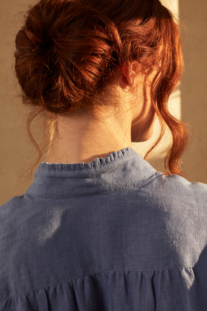 Photo en plan serré, une femme rousse se tient debout, elle est de dos avec un chignon et des mèches qui tombent sur le visage. Elle porte une chemise BOUDOIR en Lin Bleu Confectionnée dans un lin doux et léger, teinté d’un bleu ciel délicat, elle a un col officier délicatement bordé de petits volants pour une touche raffinée et ses manches légèrement bouffantes agrémentées de volants qui subliment la silhouette.