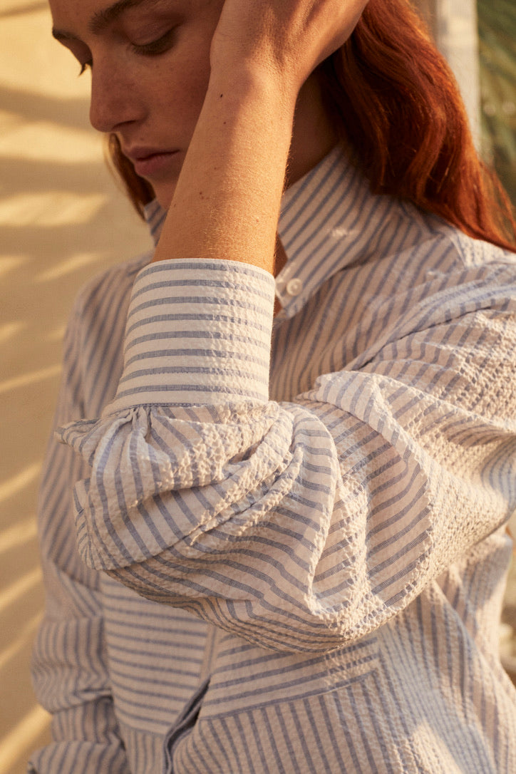 Une photo gros plan d'une femme rouse. Elle porte une chemise ENCRE SEERSUCKER, un tissu rayé bleu et blanc. Elle a un col haut et des fronces aux manches, deux grandes poches sur le devant. La femme passe sa main gauche sur ses cheveux, nous voyons bien les details du poignet. 