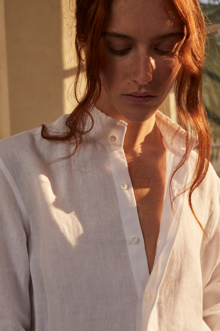 Photo plan poitrine d'une femme rousse  avec un chignon et des mèches de devant qui tombent sur son visage. Elle porte une chemise BOUDOIR en LIN Blanc aérien et léger. Des détails raffinés sur le col haut subtilement bordé de petits volants et des manches légèrement bouffantes ornées de volants, qui tombent avec élégance sur la main. 