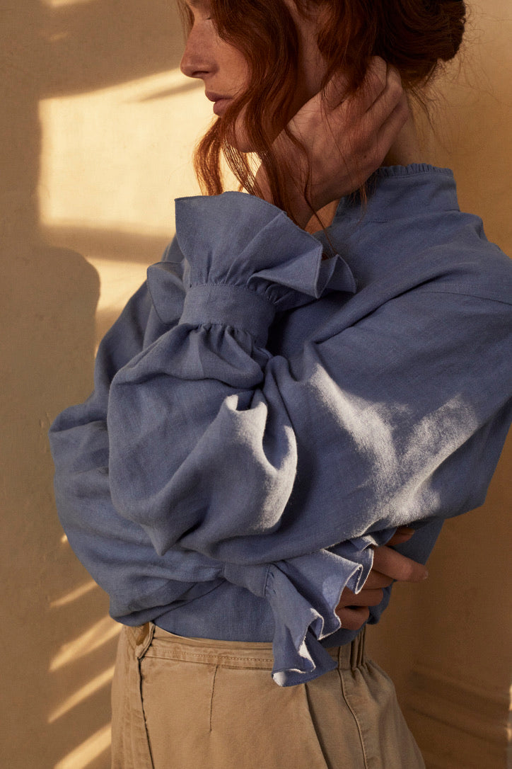 Photo en plan poitrine, une femme rousse se tient debout de profil les bras croisés avec un chignon et des mèches qui tombent sur le visage. Elle porte une chemise BOUDOIR en Lin Bleu Confectionnée dans un lin doux et léger, teinté d’un bleu ciel délicat, elle a un col officier délicatement bordé de petits volants pour une touche raffinée et ses manches légèrement bouffantes agrémentées de volants qui subliment la silhouette.