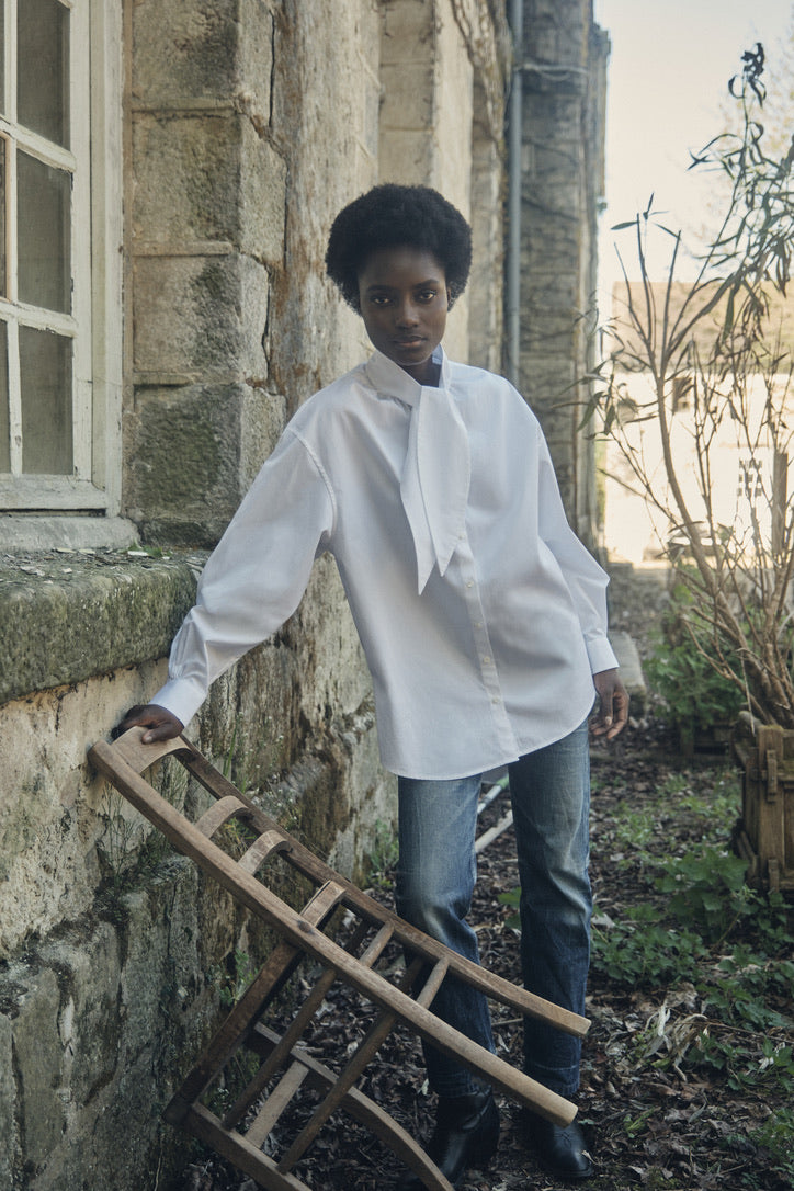 Femme portant la chemise MERVEILLEUSE, appuyée sur une chaise en bois.